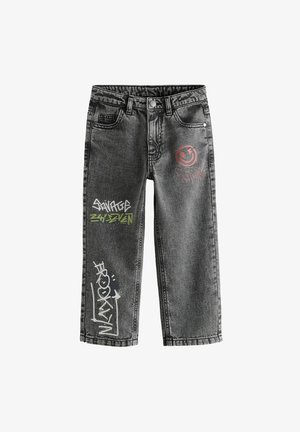 Personne portant un jean gris ample avec des motifs de style graffiti et des baskets noir et blanc, debout avec une main dans la poche.