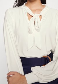 Bílá blůza s uvázanou mašlí u výstřihu, dlouhými rukávy a hladkým povrchem. U páru s námořnickým pasem s detailem hnědého pásku.