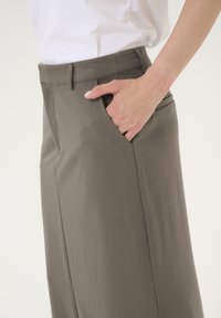 Pantalon marron clair à texture lisse, doté de poches latérales et d'une coupe ajustée, complété par un pli subtil et des passants de ceinture.