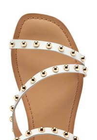 Sandalo bianco con cinturini e borchie dorate, dotato di suola piatta e di una footbed liscia color tè. Il design include dettagli cuciti lungo i bordi.