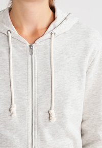 Sudadera con cremallera en un gris claro, hecha de una tela suave, que presenta una capucha con cordón y extremos anudados, y una cremallera plateada en el frente.