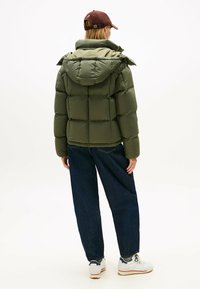 Doudoune en vert olive, avec un design boxy, une texture matelassée, une capuche, accompagnée de jean bleu et de baskets blanches.