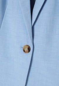 Svetlo modrý blazer vyrobený z textúrovanej látky, so zahnutým golierom a jedným okrúhlym mramorovým gombíkom na zapínaní.