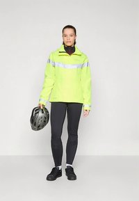Chaqueta amarilla de alta visibilidad con franjas reflectantes plateadas, combinada con leggings y zapatos negros. Sosteniendo un casco de bicicleta negro.