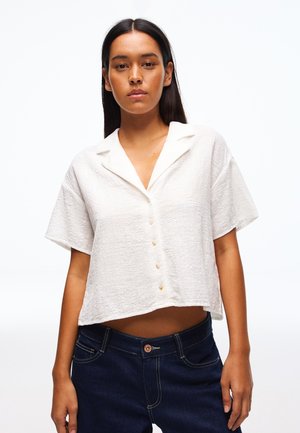 Ženska, ki nosi beli, kratkoročni teksturirani srajčni top s kratkimi rokavi, v kombinaciji s temno modrimi kavbojkami, stoji pred enobarvnim belim ozadjem.