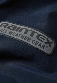 Zbliżenie na granatową tkaninę z przyszytą etykietą „Rainttex All Weather Gear” w kolorze szarym i czarnym na wodoodpornym materiale.