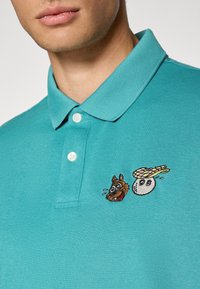 Turkis fjærgenser laget av bomull, med krage og to broderte design: en hund og en ball med en spiral, begge fargerike.