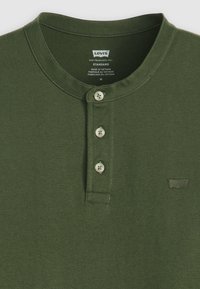 Πράσινη βαμβακερή μπλούζα henley με τρεις κουμπότρυπες και μπεζ κουμπιά; διαθέτει ένα μικρό κεντητό λογότυπο στην αριστερή πλευρά.