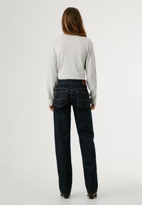 Temno modri kavbojki z visokim pasom in ravnimi hlačnicami, opremljeni z rjavim usnjenim logotipom in kontrastnim oranžnim šivom. Siva dolga majica z dolgimi rokavi.