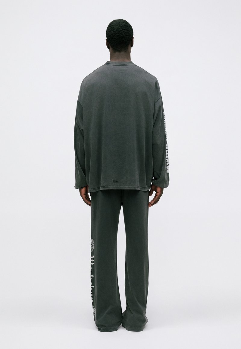 Mann mit dem Rücken zur Kamera, der ein dunkelgraues, oversized Langarmshirt und passende lockere Hose trägt, mit weißen Texten auf dem Ärmel und dem Hosenbein.