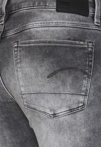 Vaqueros de mezclilla grises con una textura deslavada, que cuentan con un bolsillo trasero y una pequeña etiqueta. La costura es oscura y tiene una curva a lo largo del borde del bolsillo.