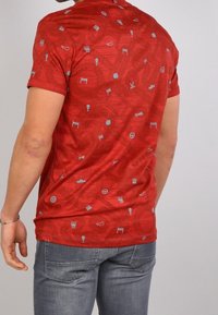 Man som bär en åtsittande röd t-shirt med små grå racingikoner, tillsammans med grå jeans, visad bakifrån mot en enkel bakgrund.