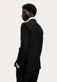 Zwart op maat gemaakt pak met een enkele split, gestructureerde schouders en een frisse witte shirt zichtbaar bij de manchet. Gladde stof met een matte afwerking.