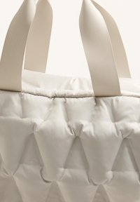 Nafouklá bílá taška typu tote s texturovanými diamantovými vzory, vybavená plochými béžovými rukojeťmi. Materiál působí měkce a vycpaně, s hladkým povrchem.