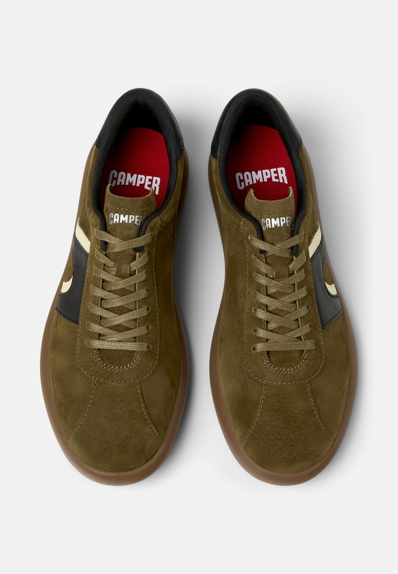 Een paar olijfgroene suède Camper sneakers met beige accenten, rubberen zolen, zwarte hielbies en rode binnenzolen met het Camper-logo.