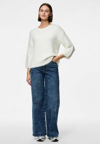 Vit fluffig tröja med vida ärmar, kombinerad med lösa blå denimjeans och silverfärgade sneakers. Bakgrunden är enfärgad vit.