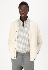 Cárdigan de punto color crema con botones en la parte delantera, combinado con una camisa a cuadros gris y blanco y pantalones grises con cordón. Estilo casual y ajuste relajado.