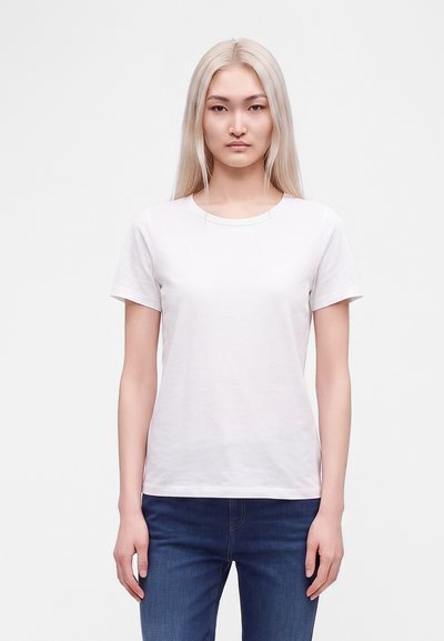 Jeune femme aux longs cheveux blonds porte un t-shirt blanc uni à manches courtes et un jean bleu, debout devant un fond blanc, face à l’avant.