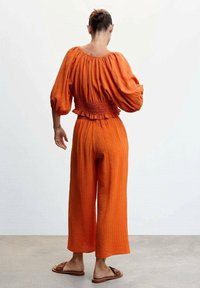 Oranžový dvoudílný outfit s texturovaným topem s puffovými rukávy a volánkovým lemem, doplněný širokými kalhotami po kotníky. Hladká tkanina, uvolněný střih.