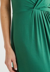 Zelená, elastická látka na šaty se zkrouceným detailem na živůtku, sklady na sukni a hladkým povrchem. Ze zblízka materiál a ruka.