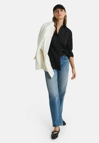 Zwarte button-up shirt, lichtblauwe jeans, witte blazer over de schouder gedrapeerd, zwarte pet en zwarte gevlochten flats. Eenvoudig, ontspannen ontwerp.