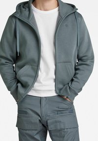 Felpa zip con cappuccio di un delicato teal, realizzata in tessuto morbido, dotata di cappuccio, due tasche frontali e cordini. Indossata sopra una maglietta bianca e pantaloni grigi.