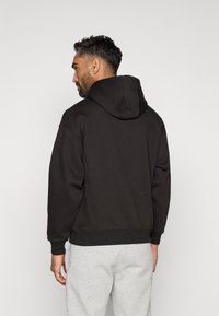 Felpa nera con cappuccio dal fit rilassato, con polsini e orlo a coste, indossata con pantaloni della tuta grigi chiari, che mette in mostra una texture del tessuto morbida.