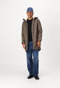 Brown padded parka with a white fleece lining, zip closure, and hood, paired with black sweater and wide-leg blue jeans. Black shoes.