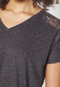 Camiseta gris oscuro con un diseño de cuello en V, que presenta detalles de encaje en los hombros y una tela ligeramente texturizada.