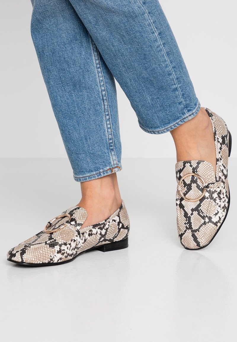 Chaussures plates à motif lézard en beige et noir avec un accent doré rond. Elles présentent une texture lisse et un petit talon, associées à un jean bleu retroussé.