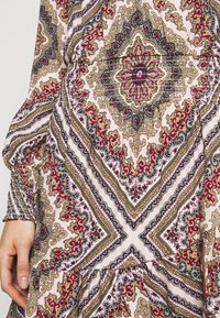 Blusa con estampado que presenta una cintura fruncida, mangas largas y un diseño con motivos florales intrincados en negro, dorado y burdeos sobre un fondo claro.