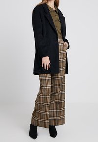 Schwarzer, strukturierter Mantel mit einem doppelt geknöpften Design, kombiniert mit braunen Karo-Hosen mit hoher Taille und schwarzen Stiefeletten.