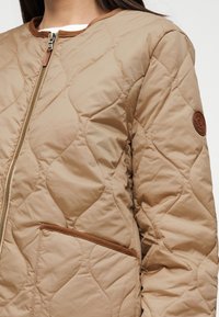Μπεζ καπιτονέ μπουφάν με λεία υφή, που διαθέτει καφέ φερμουάρ και τσέπη με καφέ λεπτομέρεια. Στρογγυλό σχέδιο για το γιακά.