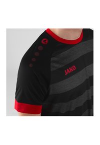 T-shirt de sport rayé noir et gris avec des accents rouges. Il présente un col rond rouge et un logo sur l'épaule, ainsi que des détails à motifs circulaires.