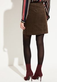 Jupe en velours côtelé marron avec poches arrière, portée avec des collants noirs et des bottines bordeaux, présentant un ourlet arrondi et une texture subtile.