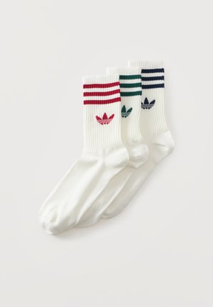 Trzy pary białych skarpet Adidas za kostkę z prążkowanymi mankietami, kolorowymi paskami i logo trefoil w kolorach czerwonym, zielonym i granatowym na gładkim tle.