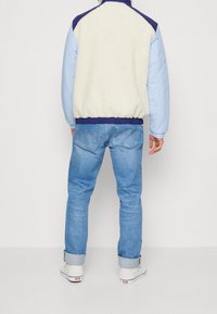 Giacca in pile e nylon, corpo crema con maniche blu, polsini e orlo a coste, indossata con jeans azzurri e scarpe bianche.