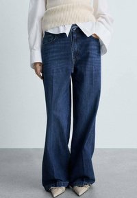 Jeans in denim blu a gamba larga con vita alta, caratterizzati da un leggero sbiadimento e tasche anteriori, abbinati a un gilet lavorato a maglia color crema e una camicia bianca.