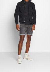 Chaqueta de mezclilla negra con botones en la parte delantera, camiseta negra, shorts grises enrollados y zapatillas altas de color claro. Fondo neutro.
