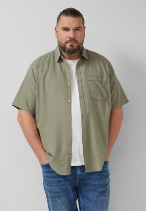 Mann mit Bart, der ein olivfarbenes kurzärmeliges Hemd über einem weißen T-Shirt und blauen Jeans trägt, steht mit den Händen in den Taschen vor einer grauen Wand.