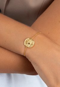 Bracciale in oro con un ciondolo rotondo e testurizzato che presenta un design a raggi di sole, attaccato a una delicata catena. Polso color pelle sottostante.