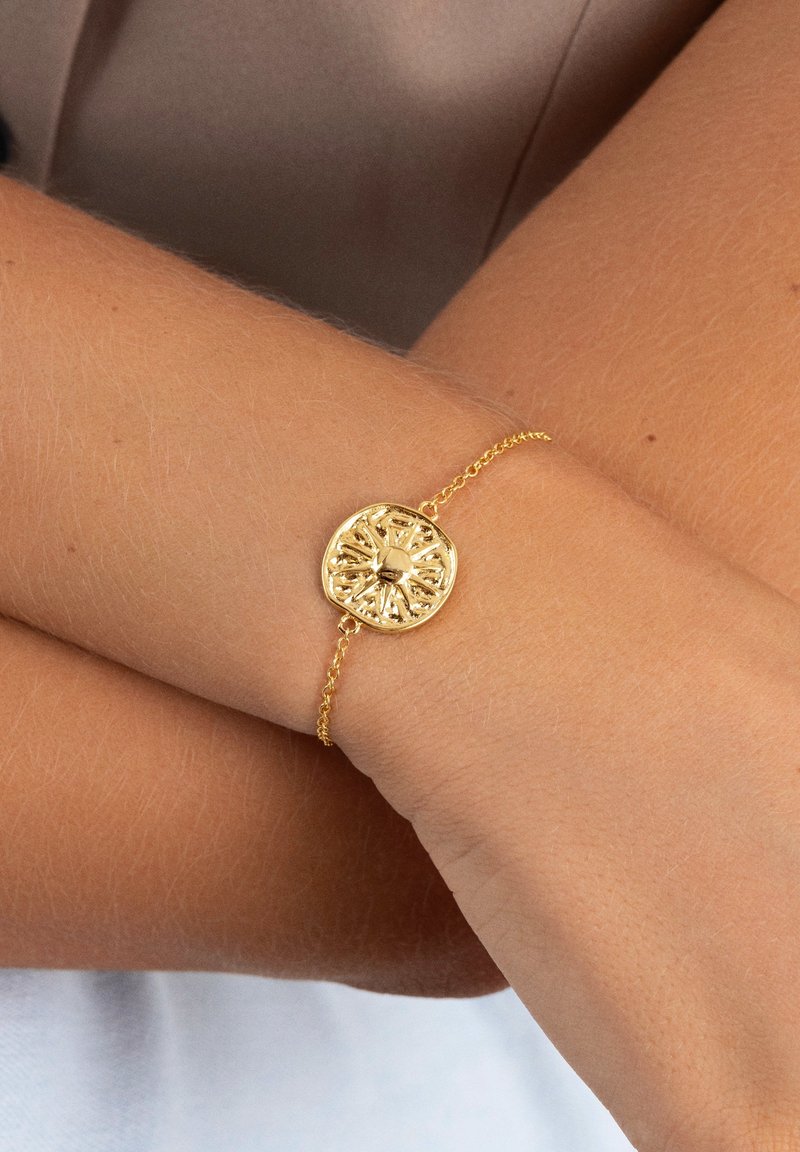 Bracciale in oro con un ciondolo rotondo e testurizzato che presenta un design a raggi di sole, attaccato a una delicata catena. Polso color pelle sottostante.