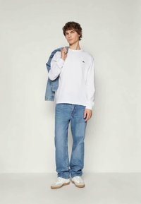 Fehér, hosszú ujjú póló kis logóval, világoskék farmernadrág és fehér sportcipő. Farmerdzseki az egyik vállán.