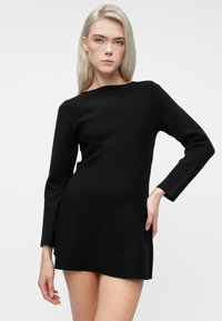 Nuori nainen, jolla on pitkät vaaleat hiukset, pukeutuneena tiukkaan mustaan pitkähihaiseen mini-mekkoon, poseeraa käsi lanteella yksinkertaista taustaa vasten.