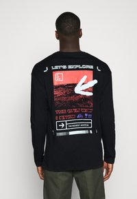 Svart långärmad t-shirt med grafiskt tryck på baksidan i rött, vitt och lila med olika texter och symboler. Tillverkad i en bomullsblandning.