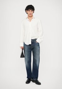 Camisa branca de mangas compridas, jeans de ganga azuis, sapatos pretos e uma pequena mala preta. Os jeans têm um lavagem ligeiramente desbotada e corte reto.