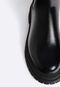 Svarta ankelboots med en blank finish, rund tå, tjock gummisula och sidodetalj med dragkedja. Slät yta och minimal sömnad.