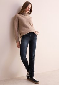 Maglione a collo alto a coste beige, jeans skinny neri e sneakers nere con lacci bianchi. Modella appoggiata a un muro neutro.