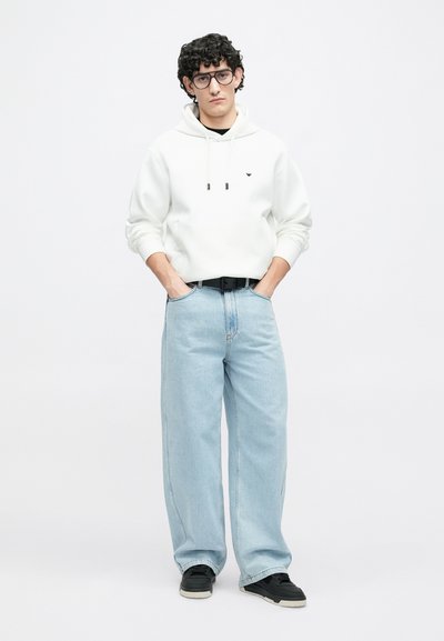 Balta su gobtuvu, su raišteliais, šviesiai mėlynos spalvos platėjantys džinsai ir juodi sportbačiai. Modelis juodais garbanotais plaukais ir su akiniais. Paprasta fono aplinka.