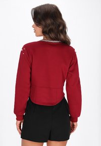 Sudadera corta roja con mangas largas, adornada con tachuelas plateadas en los hombros y una cadena decorativa en el cuello.